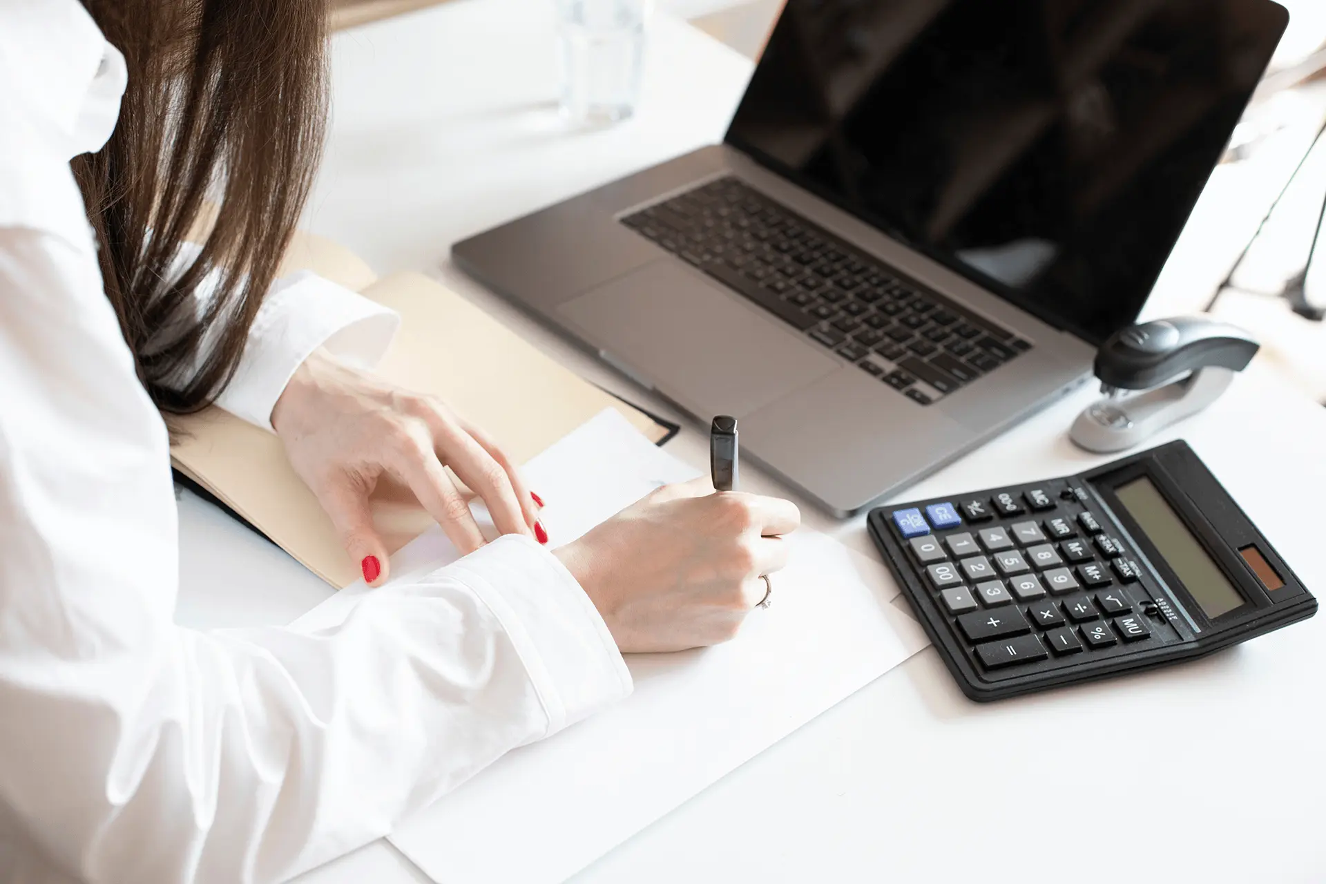 Eine Frau sitzt am Schreibtisch vor einem Laptop und einem Taschenrechner und schreibt etwas auf einen Zettel